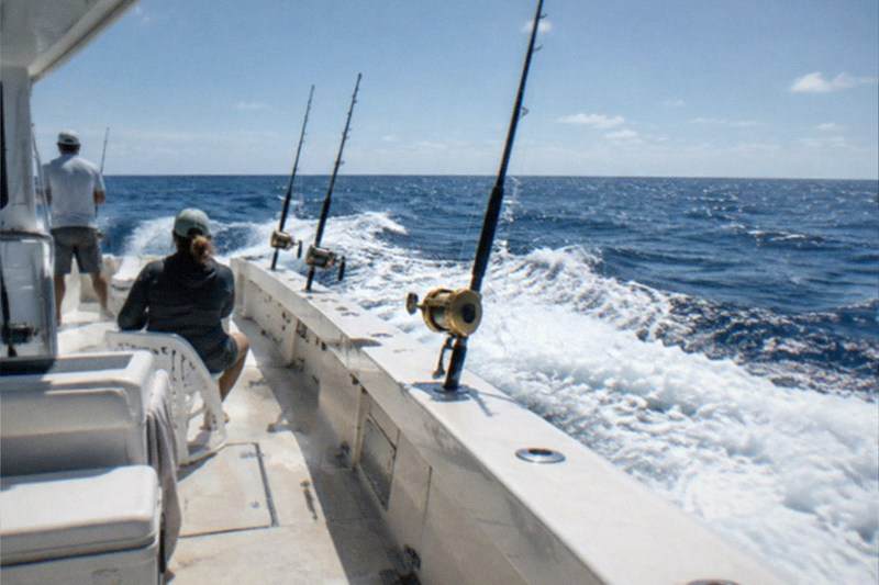 A deep sea charter boat heading out from Port Canaveral toward Atlantic offshore grounds, rods set in holders along the rail, open water beyond the inlet