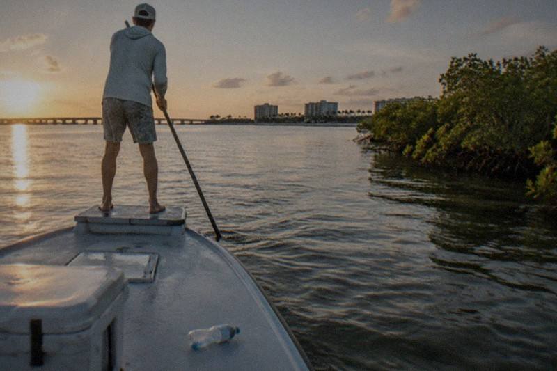 A shallow-draft skiff crossing calm lagoon flats near Cape Canaveral, mangrove shoreline behind, an angler casting toward the grass