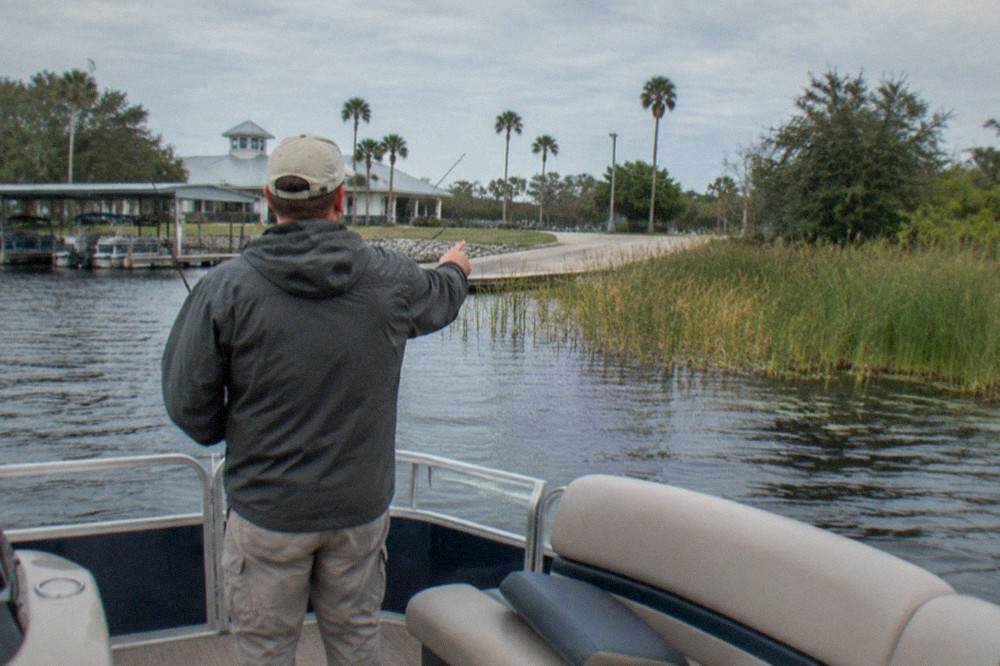 A fishing guide pointing at reeds along a lake shoreline from the deck of a bass boat