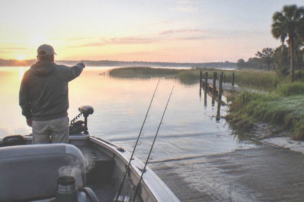 A fishing guide pointing at a spot on a lake while two rods rest against the boat rail