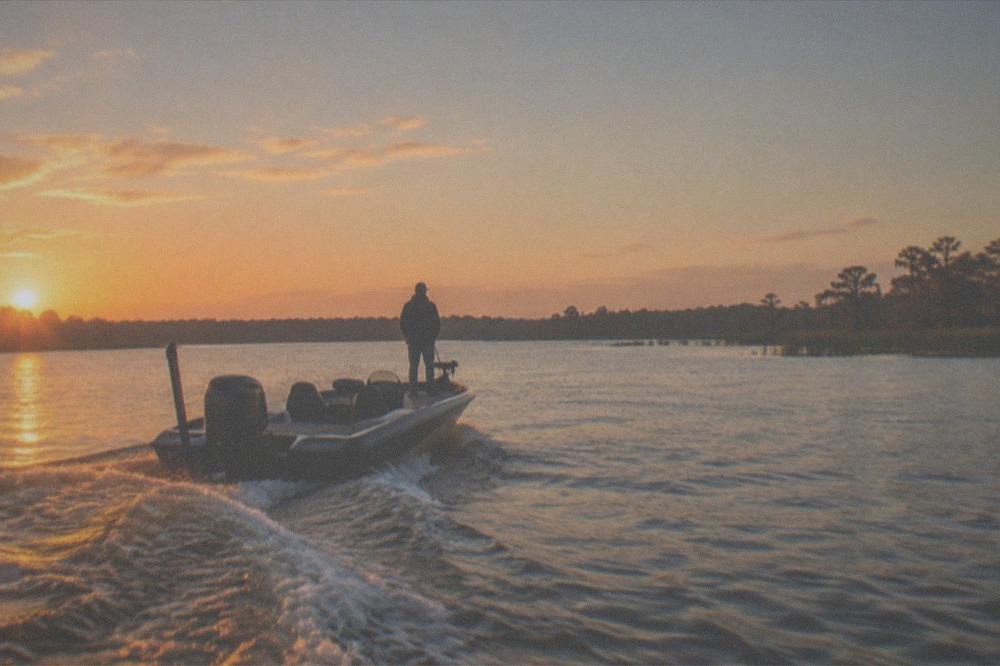 A calm Florida lake at golden hour with a bass boat motoring toward a distant shoreline of reeds